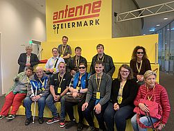 Bildtext: Gruppenfoto auf der gelben Treppe im Studio der Antenne Steiermark
